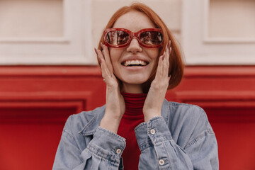 Wall Mural - Horizontal portrait of young foxy lady posing outdoors. Gorgeous smooth-haired girl with blue shirt and red sunglasses, smiling against bright background