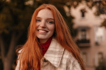 Wall Mural - Fashion portrait of joyful young ginger posing on street. Attractive girl with smooth long hairstyle, freckles and trendy outfit, smiling and looking into camera outdoors