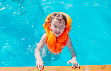 a little girl in an orange life jacket swims in the pool and looks at the camera