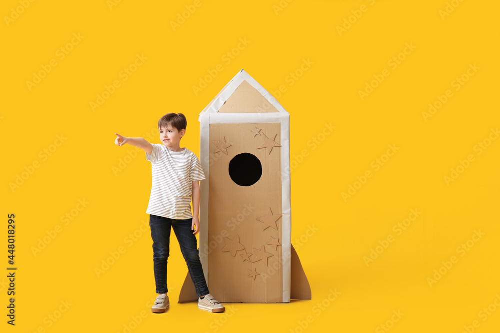 Cute little boy with cardboard rocket pointing at something on color background