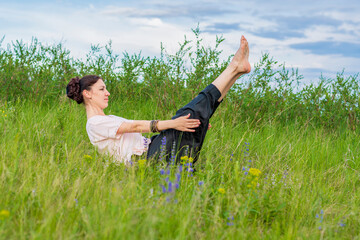 Wall Mural - Beautiful young woman practices yoga outdoors. Sits on the ground with legs raised and holds a corner.