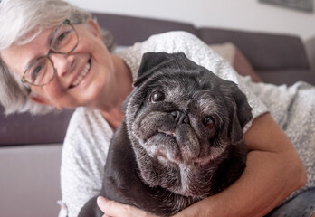 Portrait of black purebred old pug dog sitting with his senior owner on the floor at home. Best friend concept