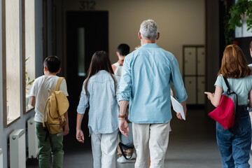 back view of teacher with digital tablet and pupils with backpacks walking along school corridor