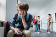 depressed boy with smartphone sitting alone in school corridor near teenagers on blurred background