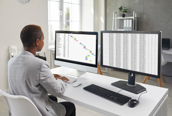 Young black woman in suit using two office computers. Financial accountant, business lady or assistant girl sitting at desk and working with data lists on modern desktop PCs. Back view, over shoulder