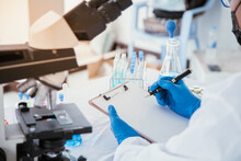 Medical Or Scientific Researcher Researching And Experimenting Multi-colored Solution, Vial And Microscope In The Laboratory Or In The Laboratory By Wearing Blue Gloves And White Clothing Completely.
