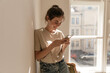 © Look! - Lifestyle portrait of young brunette looking into phone at light room. Pretty girl with collected hair and light clothes, standing against big window background and holding mobile