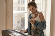 © Look! - Music, instrumental, piano, hobby concept. Stylish young lady with collected hair, white top and blue shirt, playing piano and holding phone at light room