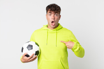 young handsome man looking shocked and surprised with mouth wide open, pointing to self and holding a soccer ball