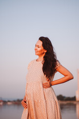 Pregnant woman against background of nature. Beautiful young Asian woman with long dark hair poses on river bank in rays of setting sun. 