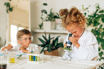 Wall Mural - Boy and girl in white uniforms and protective glasses do chemical experiments in a home laboratory.Back to school concept.Young scientists.Natural sciences.Preschool and school education of children.