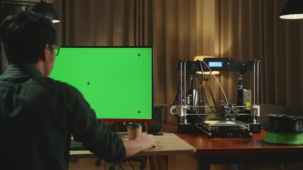 Back View Of Asian Man Work On 3D Printer And Personal Computer With Mock Up Green Screen Display In Home Office
