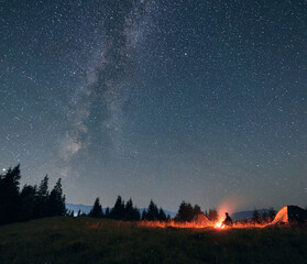 Wall Mural - male traveler looking at beautiful night sky with stars and milky way, sitting near campfire and tou