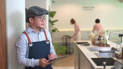 Wall Mural - Asian man coffee shop manager walking behind bar counter working on digital tablet checking and counting inventory for ordering product for cafe. Small business restaurant owner entrepreneur concept