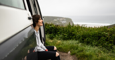 Attractive young female sitting inside a campervan while looking outside of the van and enjoying morning fresh air