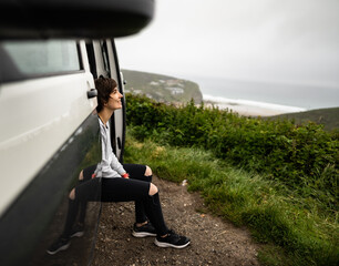 Attractive young female sitting inside a campervan while looking outside of the van and enjoying morning fresh air