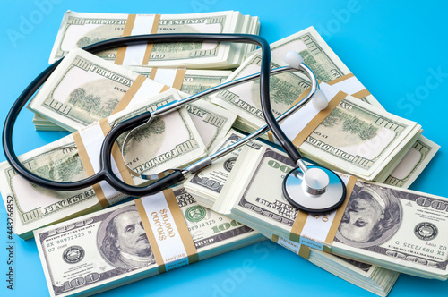 Profit driven hospital, premium medical insurance payment and high doctor bills expense concept with stethoscope, stacks of dollars and no people isolated on blue background