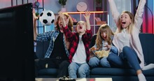 Attractive Excited Football Fans Family Sitting In Front Of TV At Home In The Evening And Rejoycing From Scored Goal With Raised Hands And Shouts