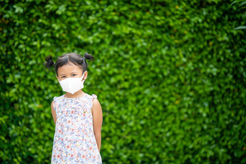 Wall Mural - Adorable small Asian girl wearing protective face mask standing against ficus annulata plant background.