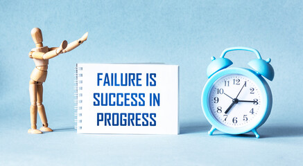 Failure is success in progress - inspirational text on a notebook, next to a wooden doll clock on a blue background.