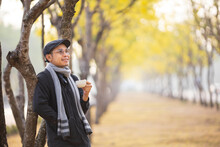 Asian Man Holding Cup Of Coffee While Walking Outdoor At The Public Park During Autumn Season While The Leaf Turning Into Yellow