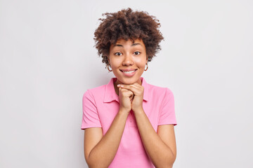 Wall Mural - Portrait of pretty dark skinned woman keeps hands under chin smiles pleasantly dressed in casual pink t shirt isolated over white background. Face without makeup. Authentic emotions concept.