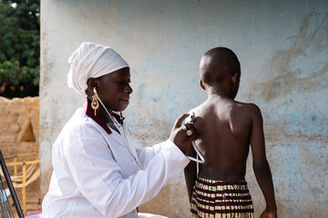 Doctor in white uniform placing a stethoscope on the back of a small black boy for measurment of respiratory sounds and heartbeat during a routine health check in West Africa