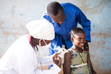 Smiling Cute African Girl Being Injected A Vaccine Dose In Her Upper Arm By A Black Female Pediatrician With Mask And White Uniform Assisted By A Blue Coated Healthcare Worker