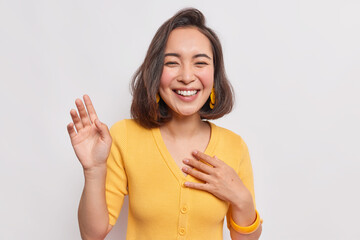 Wall Mural - Happy dark haired pretty Asian woman with positive expression laughs joyfully keeps hand raised smiles broadly wears yellow jumper earrings hears something funny isolated over white background.