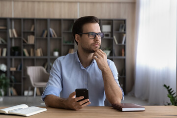 Wall Mural - dreamy young man in spectacles sit at office desk hold cellphone in hand look away of gadget screen.
