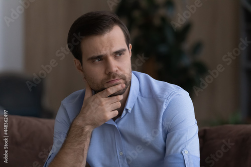 Gloomy thoughts. Head shot of unhappy upset young businessman sit on couch alone rub bearded chin look away think on debt bankruptcy losing job dismissal ponder on future life after divorce betrayal