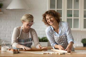 Wall Mural - Excited mature mother with grown up daughter having fun, rolling dough together, chatting laughing, cooking homemade cookies or pie, happy woman with senior mom spending leisure time at home