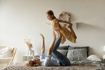 Wall Mural - Strong mother lifting adorable little daughter on legs, having fun on cozy bed at home, happy family involved in funny activity, happy adorable preschool girl and mom playing active game together