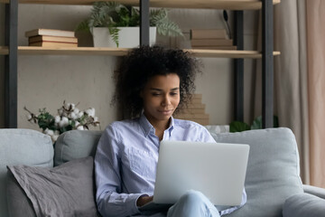 Wall Mural - Young African American woman sit relax on sofa in living room work online on laptop gadget. Biracial female look at computer screen browse wireless internet, text message on web. Technology concept.