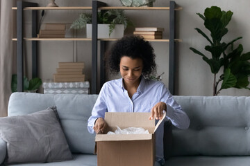 Happy millennial African American female buyer client open unpack box with internet order, buy on web. Smiling young biracial woman unbox package shop online from home. Shipment, delivery concept.