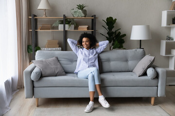 Happy young African American woman relax on couch in living room look in distance dreaming planning. Smiling biracial female renter rest on sofa at home relieve negative emotions breathe fresh air.