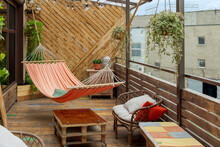 Hammock Hangs On Wooden Terrace With Table, Flower Pots And Chairs. Old Weathered Buildings In The Background. Concept Of Paradise Corner In Industrial District Of Big City.