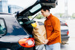 © F8  \ Suport Ukraine - Man going with cart on shopping mall parking. Young caucasian smiling male putting paper bags with food into car trunk.