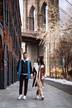 Young Caucasian Couple Of Tourists Walk At Brooklyn Bridge In New York City. Cheerful Man And Woman, Lovers On Trip, Holding Hands Happy On NYC Street. Hipsters Guy And Girl Enjoy Morning On Manhattan