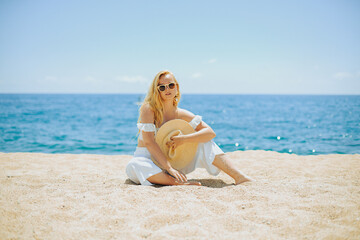 Wall Mural - A chic fashionable long-legged model in a white outfit sits on the beach against the backdrop of the ocean and enjoys life. Vacation, vacation, summer mood. Blonde in beautiful jewelry, rich woman