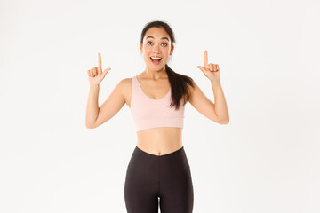 Sport, wellbeing and active lifestyle concept. Excited and happy asian fitness girl in sportswear, pointing fingers up and looking amazed with great discounts, read banner, white background