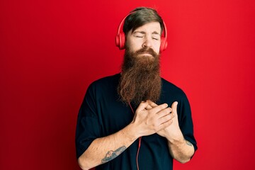 Poster - Redhead man with long beard listening to music using headphones smiling with hands on chest with closed eyes and grateful gesture on face. health concept.