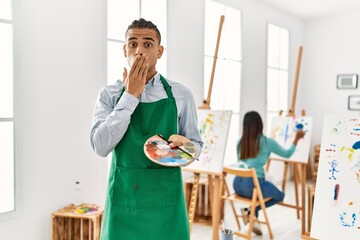 Canvas Print - Young latin man standing at art studio covering mouth with hand, shocked and afraid for mistake. surprised expression