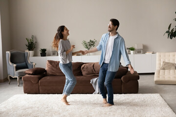 Happy excited millennial couple dancing together in new stylish cozy living room. Husband and wife celebrating buying new house, enjoying party for two, having fun on heating floor with soft carpet