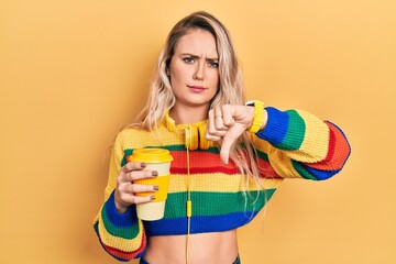 Wall Mural - Beautiful young blonde woman drinking cup of coffee wearing headphones looking unhappy and angry showing rejection and negative with thumbs down gesture. bad expression.