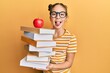 Beautiful brunette little girl holding a pile of books with an apple on the top sticking tongue out happy with funny expression.