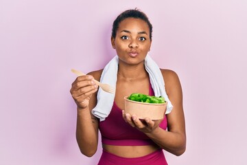 Young african american girl wearing sportswear eating salad making fish face with mouth and squinting eyes, crazy and comical.