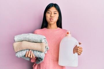 Sticker - Young chinese woman holding detergent bottle and clothes puffing cheeks with funny face. mouth inflated with air, catching air.