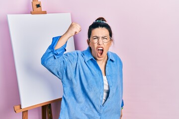 Canvas Print - Young hispanic woman standing by painter easel stand angry and mad raising fist frustrated and furious while shouting with anger. rage and aggressive concept.