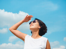 Woman In Summertime. Smiling Beautiful Asian Woman Short Hair Wearing Sunglasses And White Sleeveless Shirt Looking Up And Shading Eyes With Her Hand On Blue Sky Background On Sunny Day In Summer.